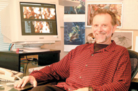 A smiling man in a red checkered shirt sits at a desk next to a computer monitor displaying images.