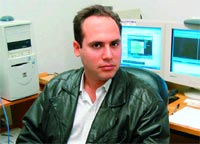 A man in a dark leather jacket and white shirt sits in an office with two computer monitors behind him.