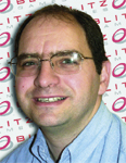 A man with short dark hair and glasses smiling against a patterned background.