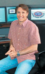 A man in a short-sleeved patterned shirt and blue jeans sits in front of several computer monitors.