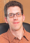 A man with short dark hair and glasses wearing an orange checkered button-down shirt.