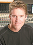 A man with light brown hair smiling in front of a large audio mixing console.