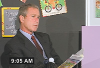 A man in a dark suit and red tie sits before a dark bulletin board with colorful papers.