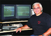 A man with gray hair and glasses smiling while sitting at a desk with two monitors and an audio mixer.