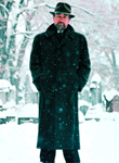 A man wearing a dark overcoat and fedora stands in a snowy, wooded landscape.