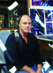 A man in a black shirt sits in a studio with colorful abstract artwork displayed behind him.