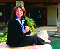 A woman wearing a black blazer and blue shirt sits on a stone ledge near a swimming pool.