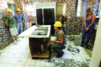 Workers in hard hats and blue overalls working in a room with patterned wallpaper and debris on the floor.
