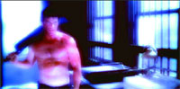 A shirtless man stands in a dark room holding a large, dark object over his shoulder near a window.
