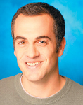 A man with short dark hair and a smile, wearing a grey crewneck shirt against a blue background.