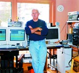 A man in a blue sweater and jeans stands with arms crossed in a room with multiple computer monitors.