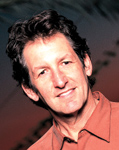 A man with dark, wavy hair smiling while wearing an orange collared shirt.