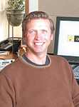 A smiling man with short brown hair wearing a brown sweater, sitting in an office setting.