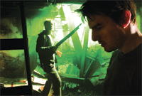 A man in the foreground looks down while another man in the background holds a long object in a green-lit room.