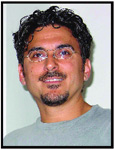 A man with dark curly hair, glasses, and a goatee smiling against a light background.