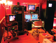 A video editing studio filled with monitors, server racks, and electronic equipment under red lighting.