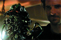A man looks at the camera while holding a dark, textured, alien-like creature with glowing red eyes.