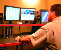 A man sits at a desk with three computer monitors, speakers, and video editing software.