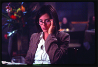 A woman with dark hair and glasses wearing a brown blazer sits at a dimly lit table.