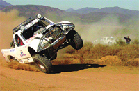A white off-road racing truck mid-air, tilting heavily while kicking up dust on a dirt track.