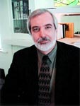 A man with graying hair and a beard wearing a dark blazer, patterned tie, and collared shirt.