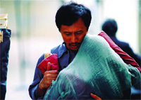 A man in a blue shirt carries a large teal bundle and a red bag.