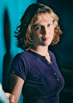 A woman with wavy blonde hair wearing a purple short-sleeved shirt looks forward.