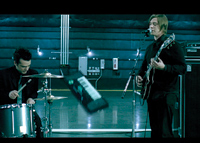 A drummer playing a snare drum and a guitarist playing an electric guitar in a dimly lit studio.