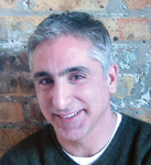 A smiling man with short gray hair wearing a dark sweater against a brick wall.