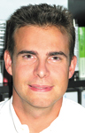 A close-up portrait of a man with short brown hair and a slight smile, wearing a white shirt.
