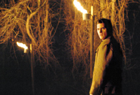 A man in a jacket looks toward the camera amidst vines and lit torches at night.