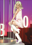 A blonde woman in a pink dress and white boots poses against vertical poles and a purple background.