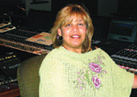 A smiling woman with blonde hair wearing a light green embroidered top in a music studio.
