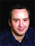 A man with short dark hair and light stubble wearing a dark blue button-down shirt against a black background.