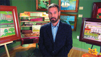 A man in a dark blazer and blue shirt stands in a room filled with various framed landscape paintings.