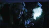A blonde woman looks intently at a bright, glowing blue light held in her hands in a dark setting.