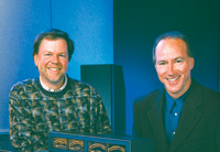 Two men smiling, one in a patterned sweater and one in a dark blazer and blue shirt.