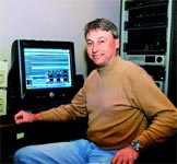 A man in a tan turtleneck sits at a desk next to a computer monitor and a rack of electronic equipment.
