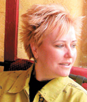 A woman with short, spiky blonde hair wearing a yellow shirt and looking toward the right.