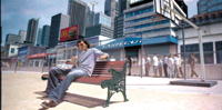 A man sits on a wooden bench in a city plaza with skyscrapers and a basketball hoop in the background.
