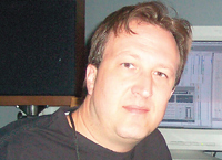 A man in a black shirt looking toward the camera in front of a computer monitor.