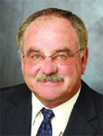 A man with short graying hair, a mustache, and glasses wearing a dark suit and patterned tie.