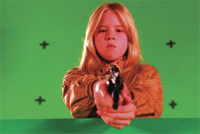 A young girl with blonde hair points a handgun directly at the camera against a green screen background.