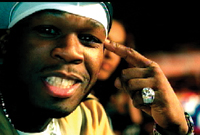A man in a white head covering smiles and points a finger to his temple while wearing a large ring.
