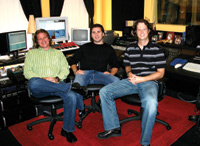 Three men sitting in office chairs on a red rug in a studio filled with computer monitors and audio equipment.