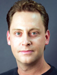 A headshot of a man with wavy brown hair and light-colored eyes looking directly at the camera.