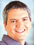 A man with short, salt-and-pepper hair smiling at the camera against a light blue background.