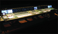 A wide professional audio mixing console with numerous sliders, knobs, and integrated digital screens in a dark room.