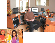 A man sits in an office chair behind a large wooden desk equipped with audio mixing consoles and computer monitors.