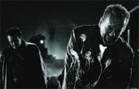 Black and white film still of two men in dark clothing standing in a dimly lit, smoky environment.